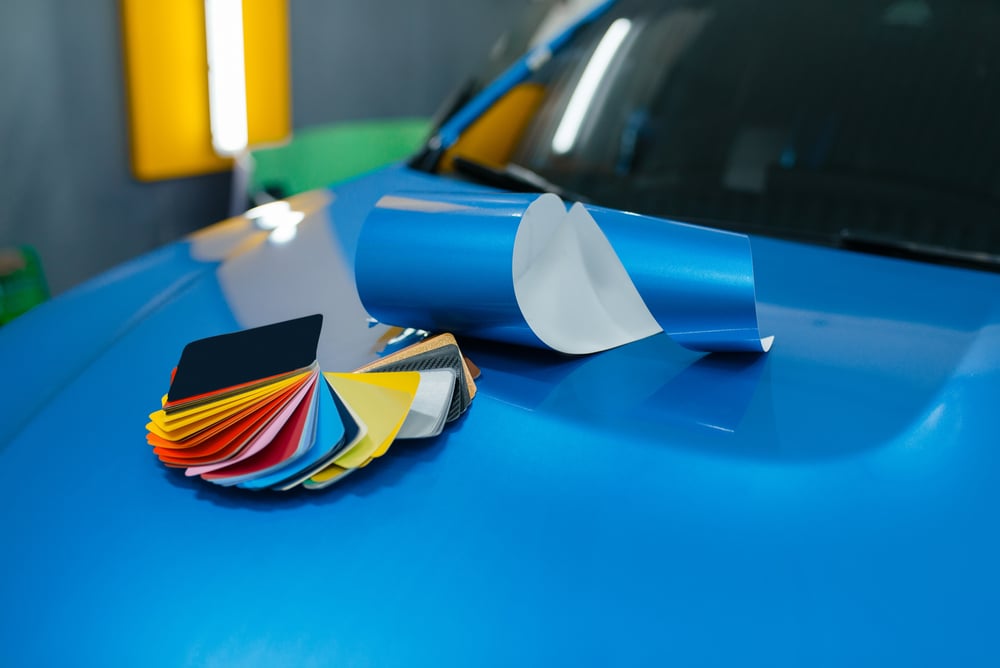A roll of blue vinyl car wrap and a swatch book of various wrap colors and finishes sitting on a glossy blue car hood.