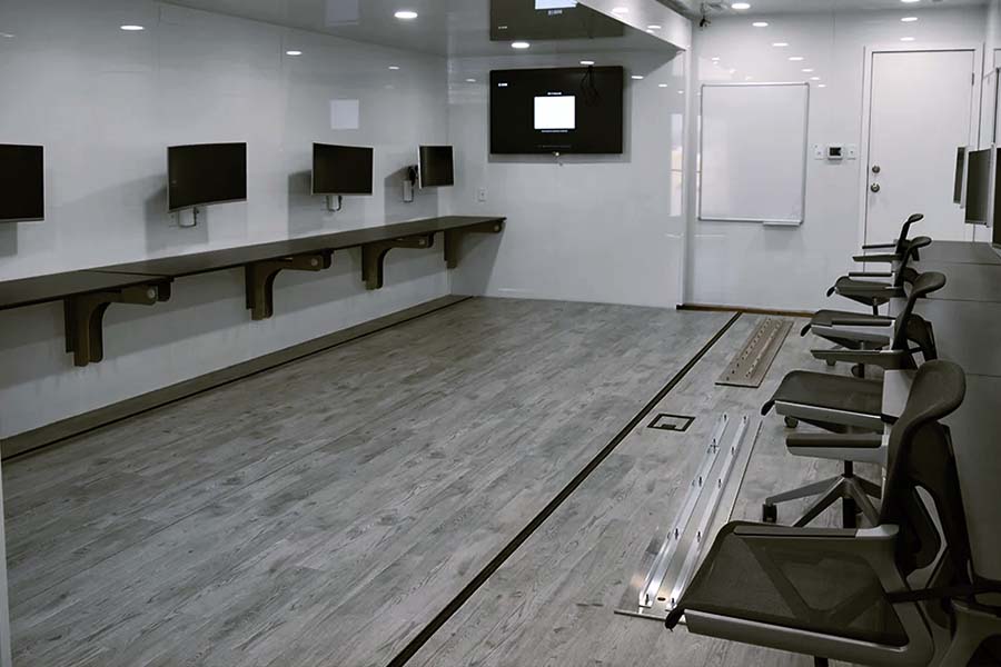 Interior of a modern mobile command or training center with wood-look flooring and two rows of black desks and monitors along the walls. The room includes a large wall-mounted television, a whiteboard, and rolling office chairs.