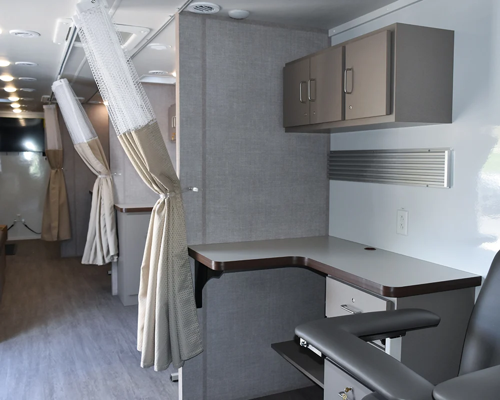 Interior of an all-inclusive mobile medical unit showing separate patient stations with privacy curtains, built-in cabinets, a long countertop desk, and a phlebotomy or medical care chair.