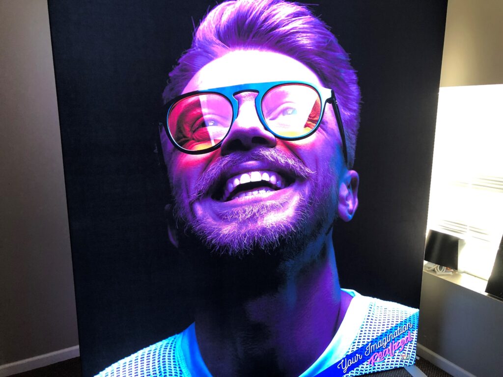 Large backlit fabric display graphic featuring a close-up of a smiling man with purple lighting and retro sunglasses.