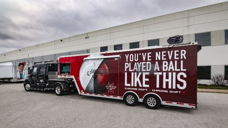 Black semi-truck hauling a dark red Callaway Golf Chrome Soft experiential marketing trailer, featuring a large golf ball graphic and the bold slogan, 'You've never played a ball like this'.
