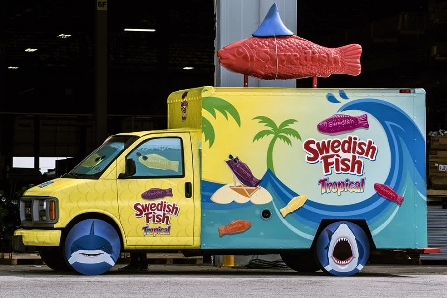 Close-up side view of the bright yellow and blue Swedish Fish Tropical promotional box truck, featuring a large 3D red fish on the roof, palm tree graphics, and playful shark-themed wheel covers for experiential consumer marketing.