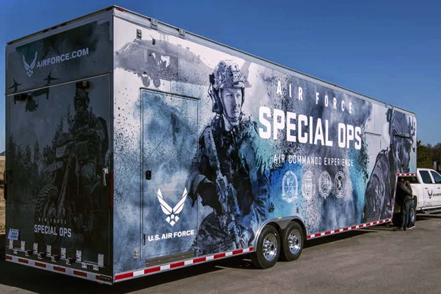 Exterior of a large U.S. Air Force Special Ops promotional trailer with a full-color graphic wrap featuring airmen in combat gear and various military insignia. The "Air Commandos Experience" trailer is parked on pavement with a tow vehicle attached.