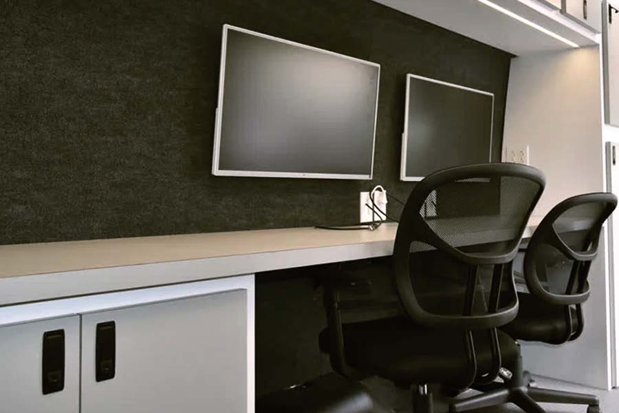 Interior workstation of a mobile command center, showing a long white desk, two black mesh office chairs, two wall-mounted computer monitors, and white storage cabinets underneath.