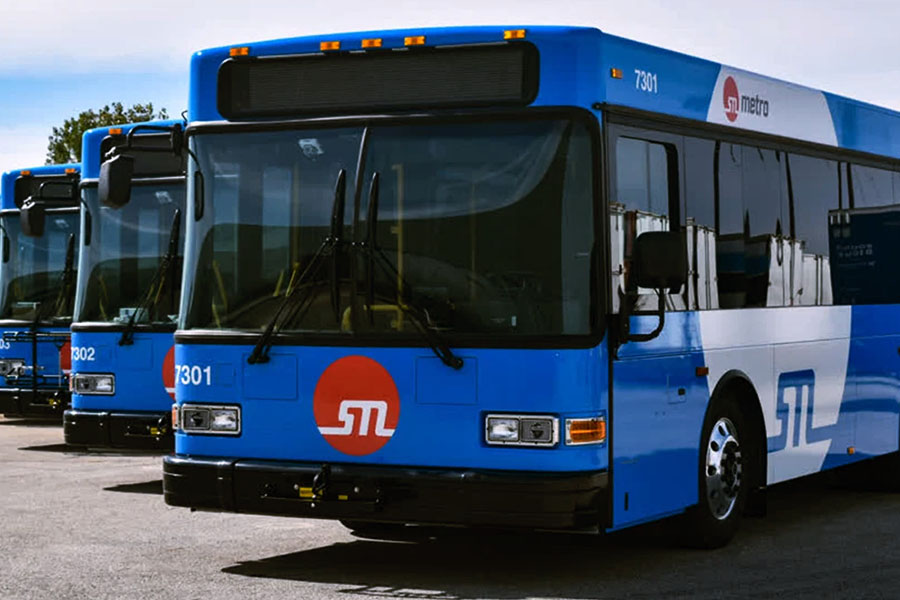 A line of St. Louis Metro public transit buses with blue and white fleet graphics are parked in a depot on a sunny day.