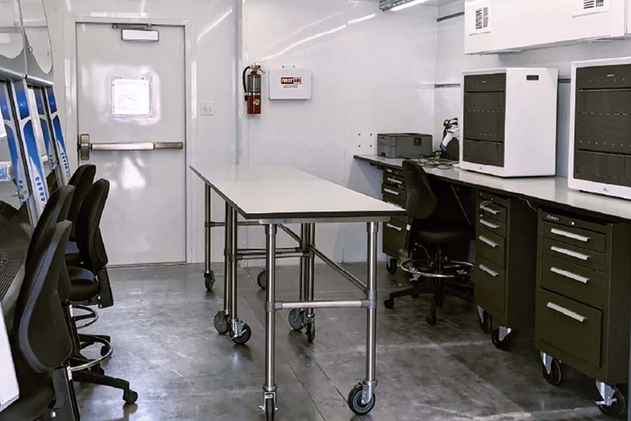 Interior of a mobile command center or office trailer featuring a long, white table on wheels, multiple computer workstations, and dark metal storage cabinets. A fire extinguisher and first aid kit are visible near the rear exit door.