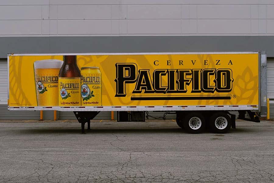 Side view of a yellow Cerveza PacĆfico beer trailer wrap featuring the large Pacifico logo and graphics of a glass, bottle, and can of Pacifico Clara beer.