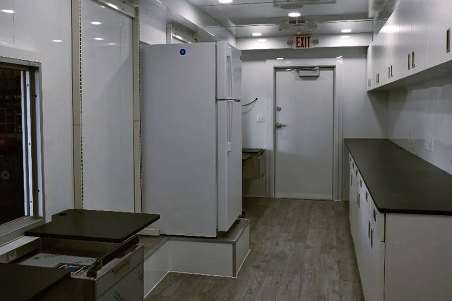 Interior of a mobile medical lab trailer featuring a commercial refrigerator, long black countertops, white cabinets, wood-look flooring, and an emergency exit door.