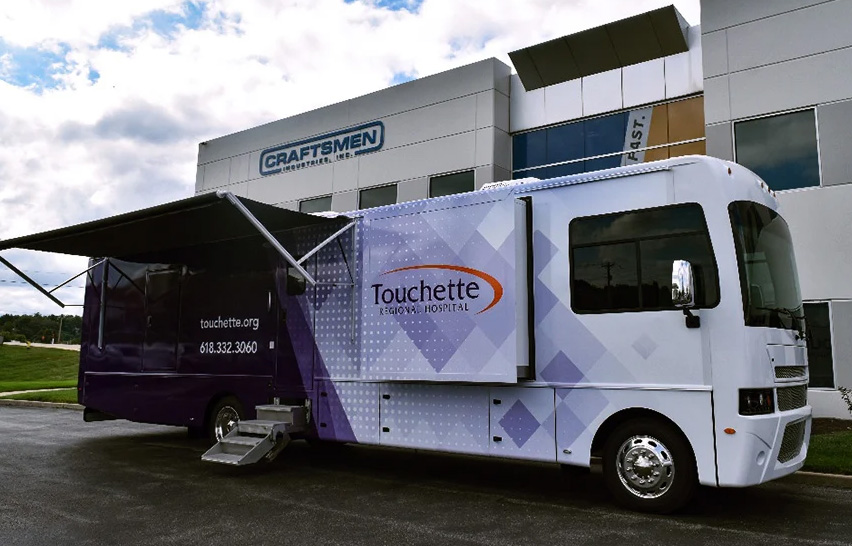 Touchette Regional Hospital mobile medical vehicle, identifiable as a custom Medical Coach-vs-Medical Bus hybrid design, featuring purple and white branding, an extended awning, and a deployable entrance staircase for patient outreach.