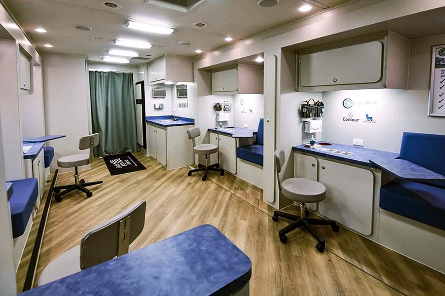 The interior of a clean clinic, with multiple exam stations and seating areas. This transportable health unit has wood-style flooring and light blue countertops with white cabinetry.