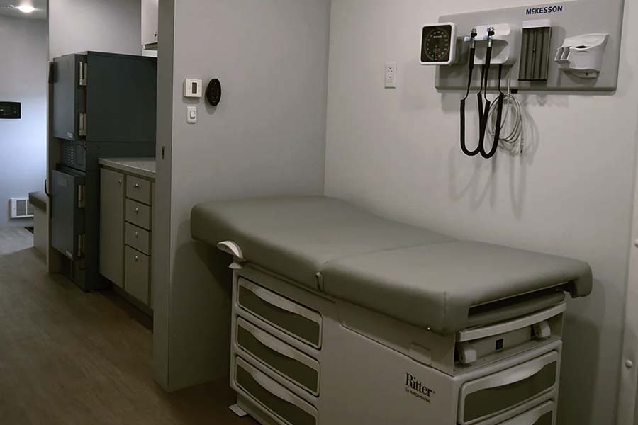 Interior of a small, well-equipped mobile clinic examination room featuring a Ritter exam table with drawers and a wall-mounted McKesson vital signs unit. The space also includes cabinetry and medical storage.