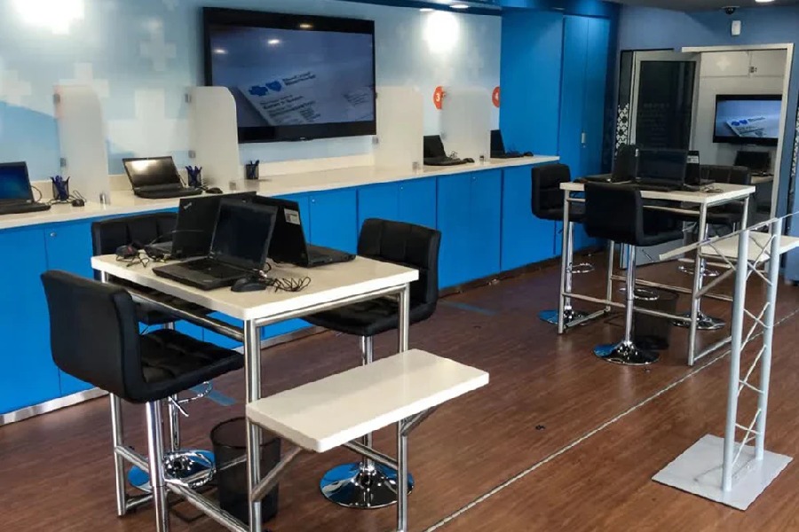 Interior of a mobile health and wellness trailer, showing a modern blue and white color scheme, high-top tables with black laptops, and a large monitor on the back wall.