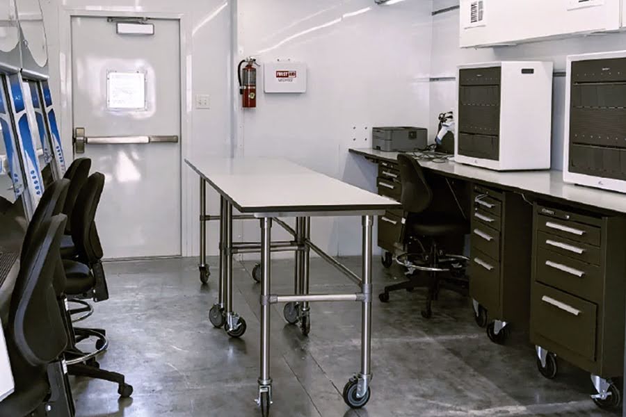 Interior of a mobile medical lab with a long stainless steel mobile work table on casters, black rolling chairs, and integrated lab equipment on fixed countertops with storage drawers.