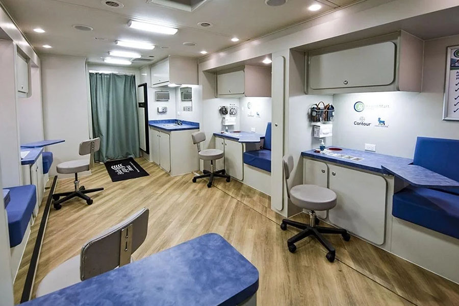 The interior of a clean clinic, with multiple exam stations and seating areas. This transportable health unit has wood-style flooring and light blue countertops with white cabinetry.