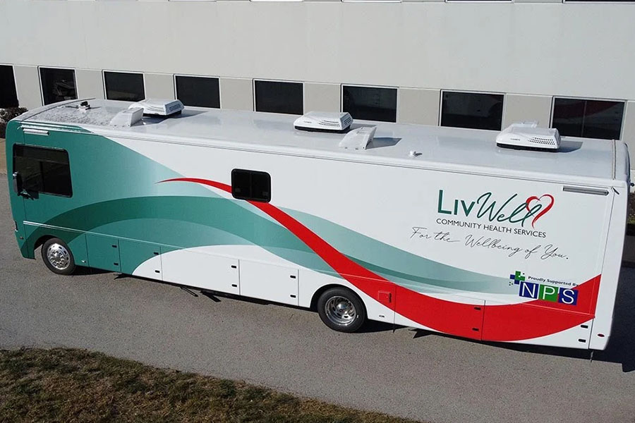 Exterior of a large LivWell Community Health Services mobile clinic motorhome with a white body and a flowing red and teal graphic design. The vehicle has multiple rooftop HVAC units, indicating an expansive interior for medical services.