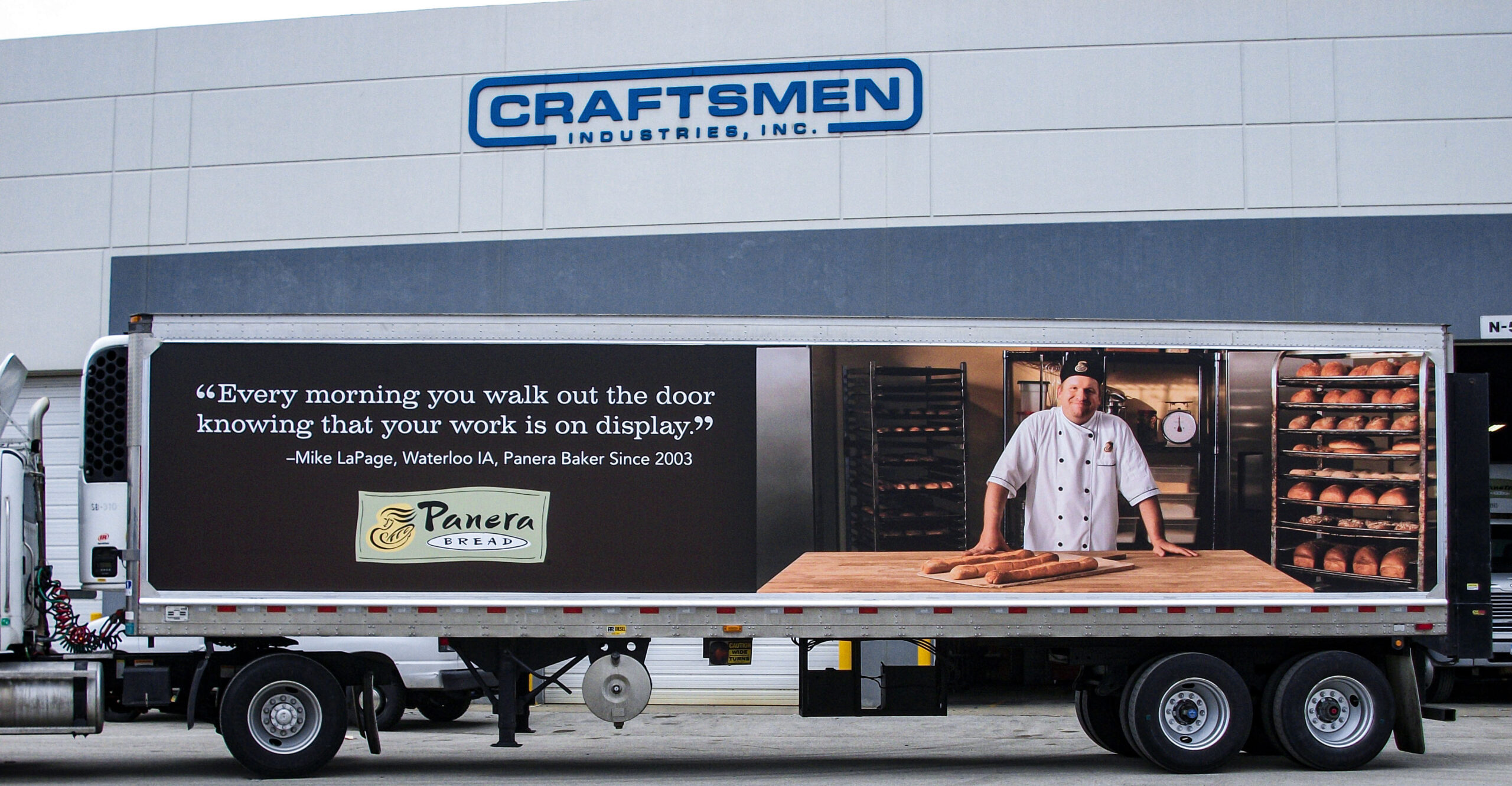 Side of a refrigerated semi-trailer wrapped with a large Panera Bread graphic featuring a baker, Mike LaPage, with loaves of bread. The trailer is parked outside the Craftsmen Industries building.