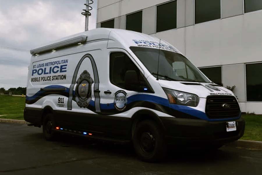 Exterior of a St. Louis Metropolitan Police Mobile Police Station van with white, black, and blue graphics, including the 911 number and city seal. The police van features a roof-mounted antenna and is parked outside a modern building.