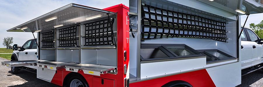 Side view of a red and white mobile service or disaster relief truck with a large, open side panel revealing custom storage compartments, black cargo netting, and two top-loading freezers or bins.