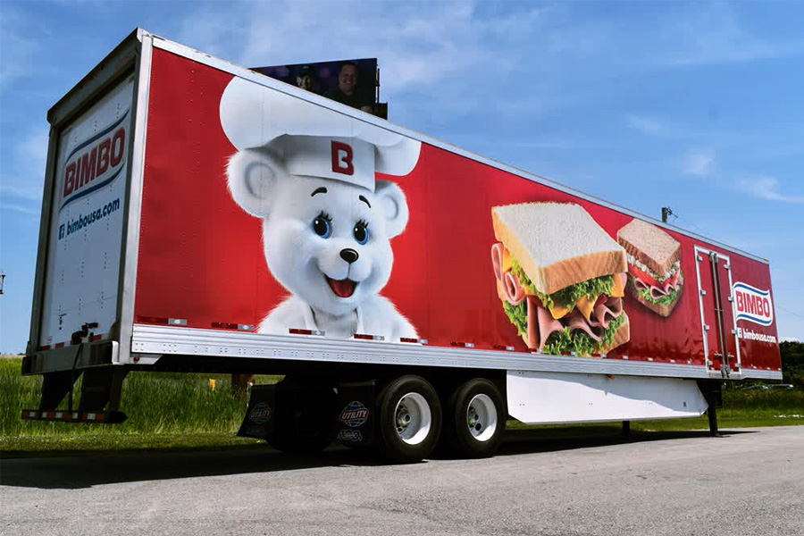 Large semi-trailer fleet graphic wrap for Bimbo Bakeries, featuring a vibrant red background, the friendly Bimbo Bear mascot wearing a chef's hat, and high-resolution images of fresh, stacked deli sandwiches to promote food product quality and brand recognition.