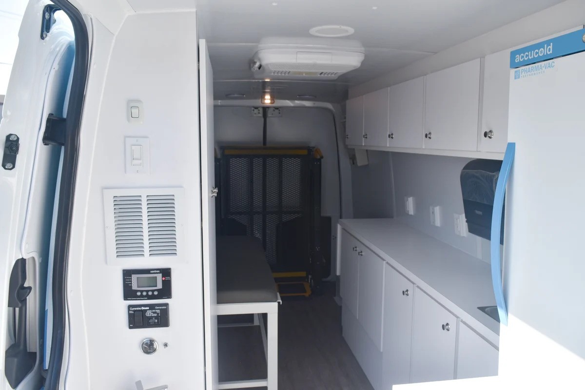Interior of a white mobile medical or vaccine van showing essential required equipment including built-in storage cabinets, a PHARMA-VAC medical refrigerator, a small exam bench, and a wheelchair lift/ramp in the rear.