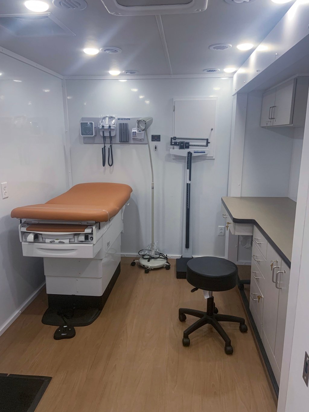 Interior of a mobile medical exam room with a padded exam table, medical scale, wall-mounted diagnostic equipment, wood-look flooring, and white cabinetry, designed for patient privacy.