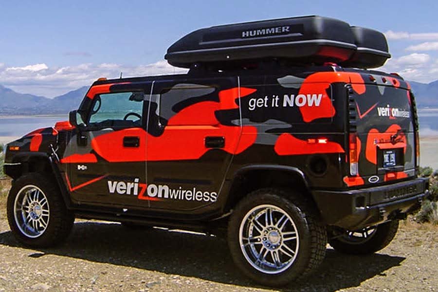 A custom Verizon Wireless promotional Hummer H2 SUV with a black, red, and gray camouflage wrap and a rooftop cargo box. The vehicle is parked outdoors with a lake and mountains in the background.