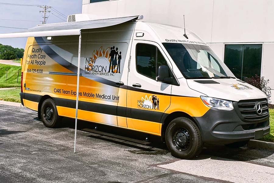 Exterior of a New Horizon Family Health Services mobile medical van parked outside an office building. The van, wrapped in a white and yellow design, has an awning extended.