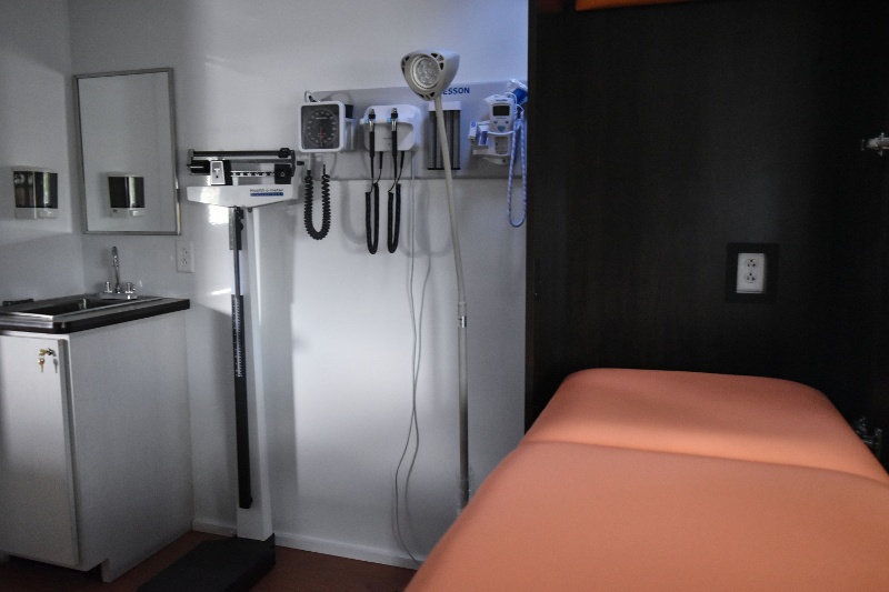 Interior of a Mobile Medical Unit examination room featuring a scale, wall-mounted diagnostic tools for blood pressure and vital signs, a sink, and a cushioned exam bed, set up for comprehensive mobile primary care.