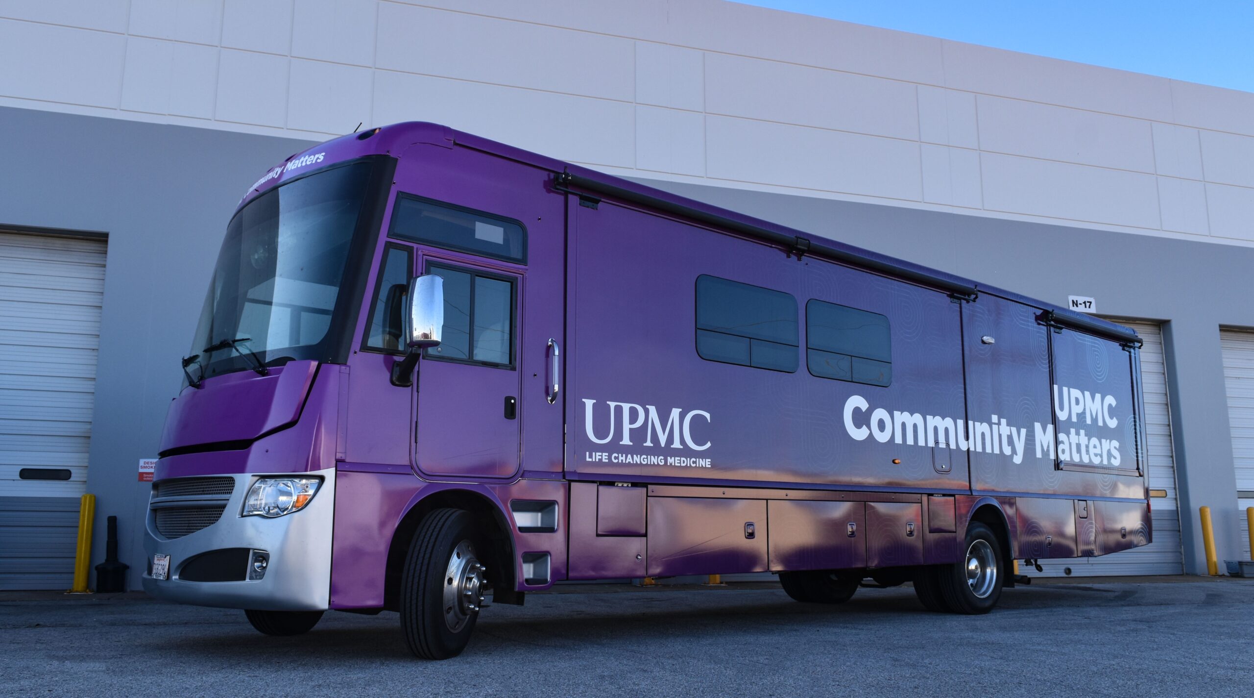 Large, bright purple UPMC Community Matters mobile medical coach with silver accents, parked outside a warehouse with the UPMC 'Life Changing Medicine' logo visible.