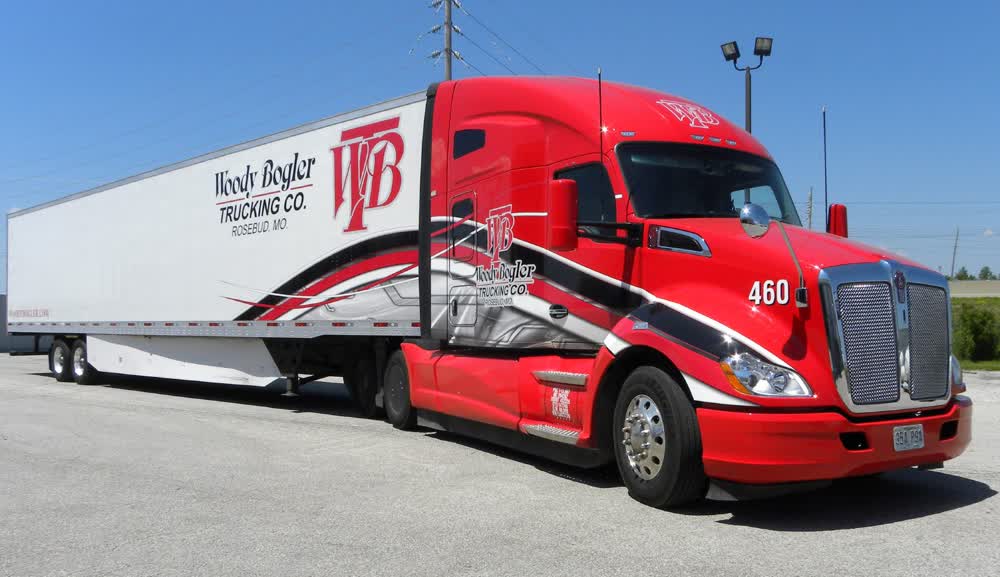 Bright red Woody Bogler Trucking Co. semi-truck cab wrap with a dynamic black and white swoosh graphic that extends onto the white cargo trailer, showing the 'WB' logo.