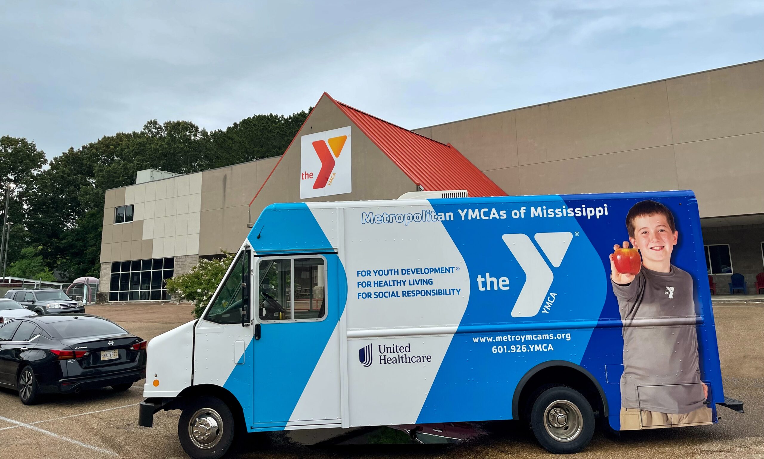 White step van with a vibrant blue and white YMCA of Mississippi vehicle wrap, featuring a young boy holding an apple and branding for United Healthcare, parked in front of a YMCA building.