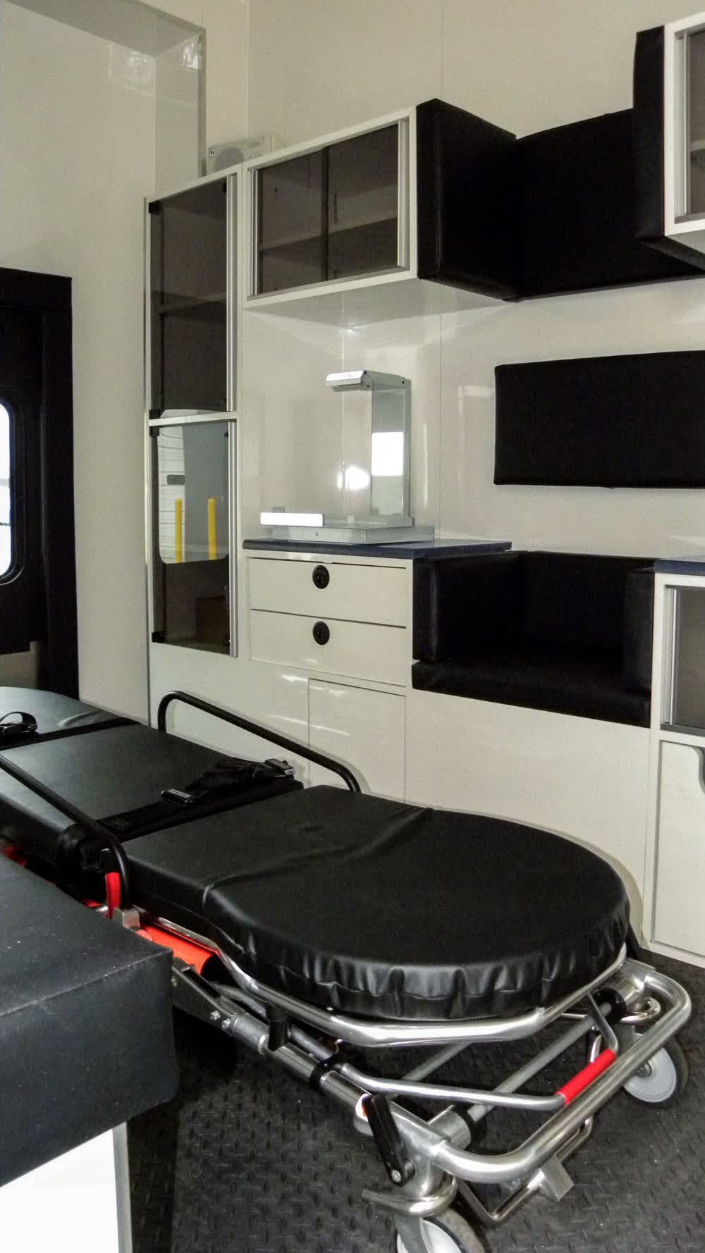 Interior of a mobile medical trailer showing a black patient transport stretcher or medical bed secured near white cabinets with black fronts, designed for emergency and field patient care.