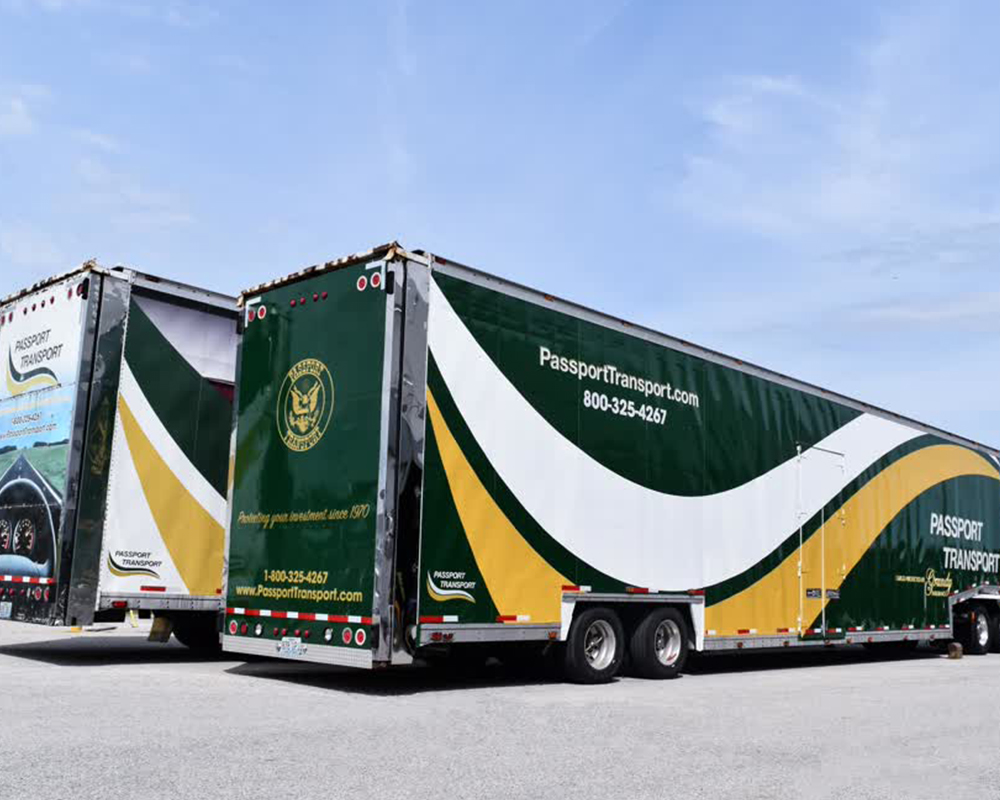 Two Passport Transport enclosed car hauler trailers with custom green, gold, and white fleet graphics. The trailers advertise the company's phone number and feature a large, sweeping logo design.