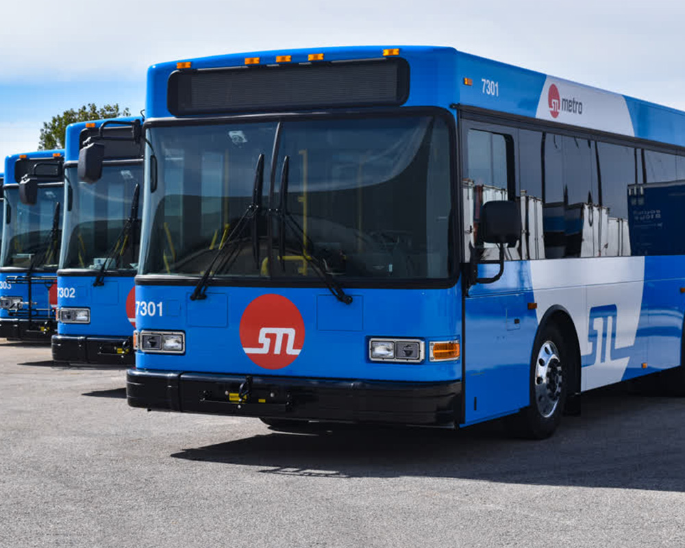 A line of St. Louis Metro public transit buses with blue and white fleet graphics are parked in a depot on a sunny day.
