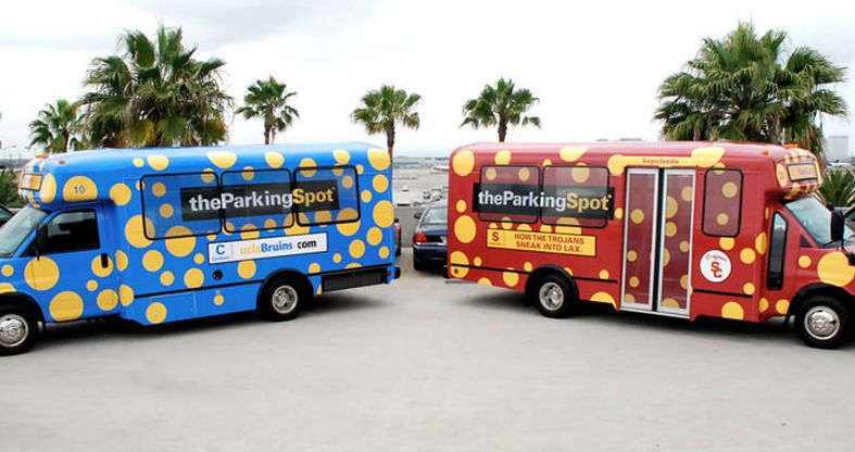 Two small, custom-wrapped airport shuttle buses for The Parking Spot, one blue and yellow for UCLA and one red and gold for USC, are parked side-by-side. The vehicles' graphics feature large circular patterns and palm trees are visible in the background.