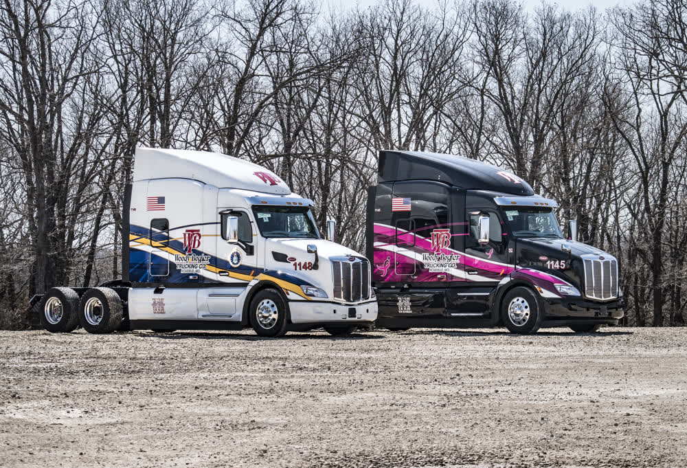Two custom-wrapped semi-truck tractors for Wolf Baylor Trucking Co., one white with blue and gold graphics and one black with pink and white graphics, are parked side-by-side.