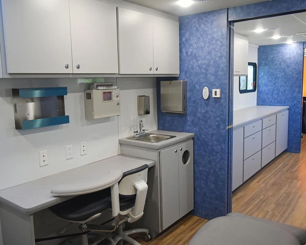 Interior of a mobile dental vehicle showing a hallway with a blue textured wall and a large mirror reflecting the front cabin area with dental chairs and a printer on a counter.