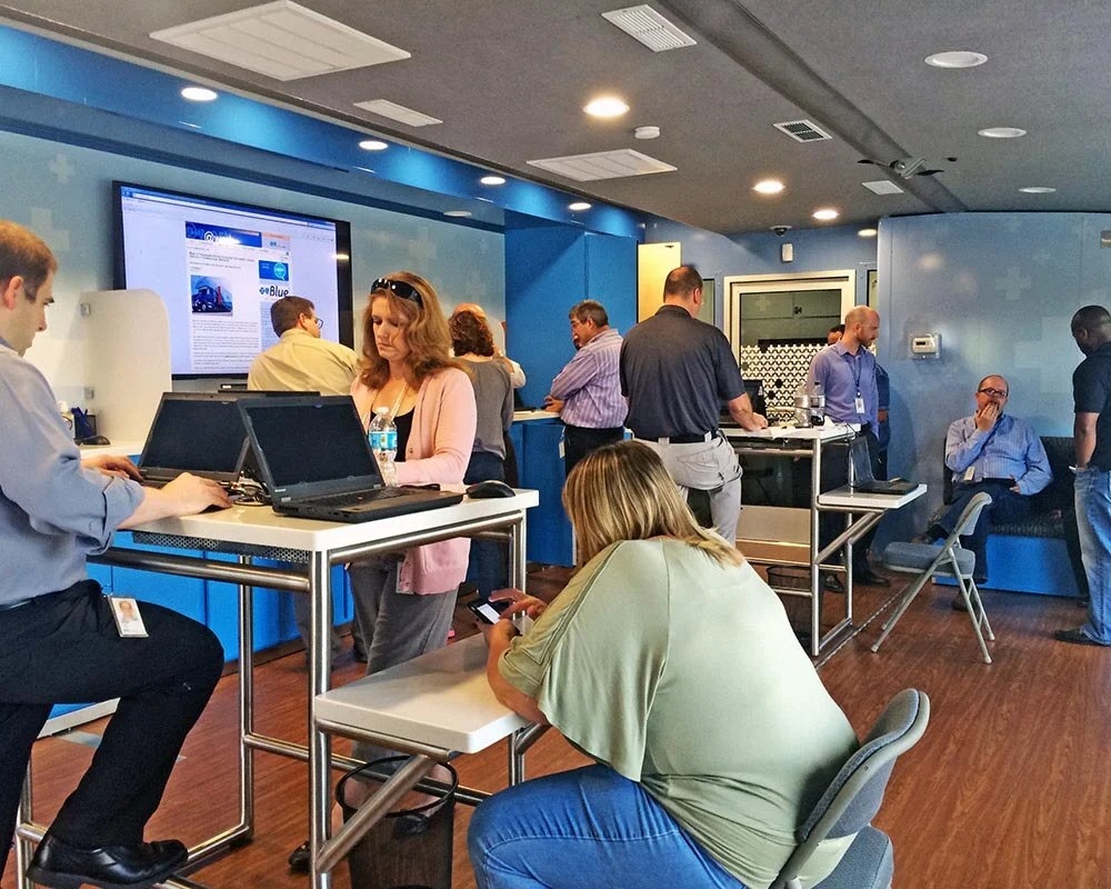 Busy interior of a BCBS 2 mobile unit or event activation with staff and visitors working at high white tables, blue walls, a large TV screen, and a dark wood-look floor.