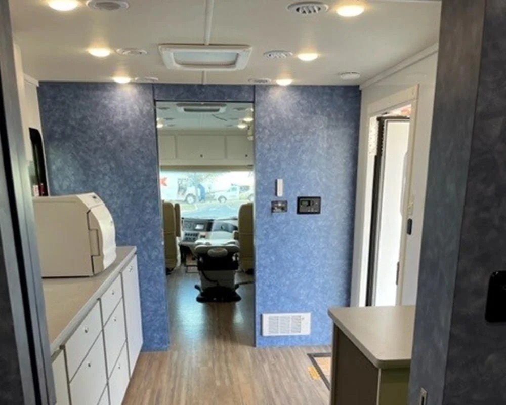 Interior of a mobile dental vehicle showing a hallway with a blue textured wall and a large mirror reflecting the front cabin area with dental chairs and a printer on a counter.