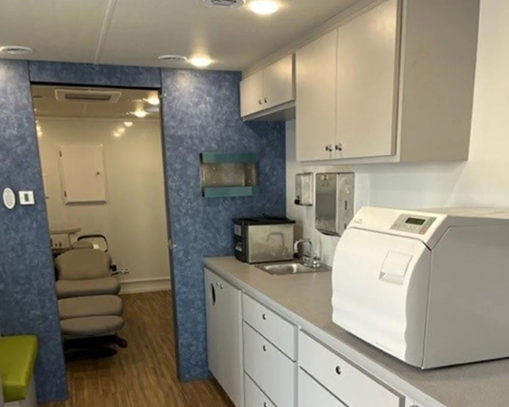 The interior of a mobile dental vehicle featuring a counter with a sink, sterilization equipment, and overhead cabinets. A dental chair is visible in the adjacent exam room.