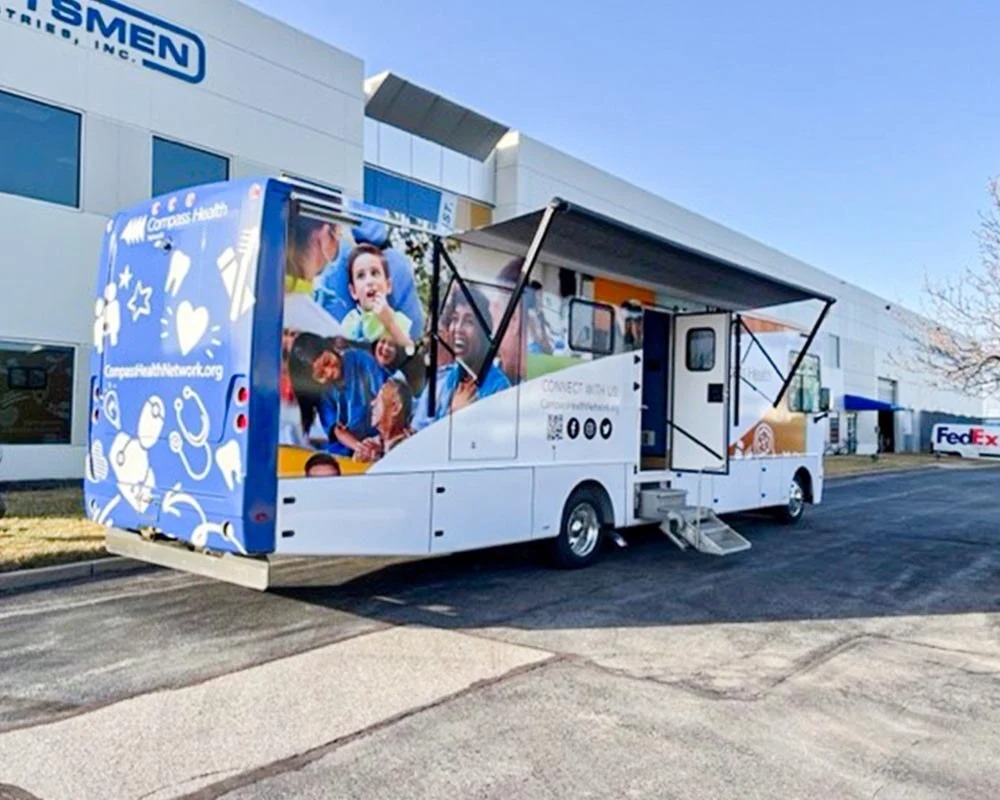 Compass Health dental trailer parked with an awning extended, a full-length graphic wrap depicting diverse smiling people, and a blue rear panel with dental-themed icons.