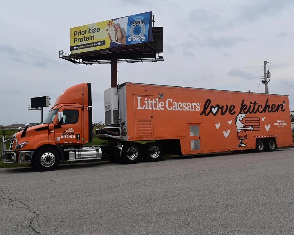 External view of a large orange Little Caesars Love Kitchen semi-truck and trailer parked under a billboard.