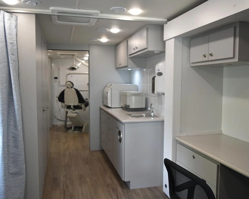Interior of a mobile dental vehicle showing a hallway with a countertop, a sink, sterilization equipment, and a view into a private dental operatory with a dental chair.