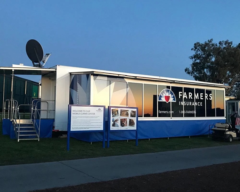 Exterior of the Farmers Onsite-1 Mobile Claims Center trailer at dusk, a large white and blue unit with the Farmers Insurance logo and a satellite dish on top, set up on a grassy field.
