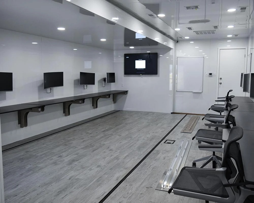 Interior view of a modern mobile command center with multiple mounted computer monitors, black office chairs, and whiteboards.