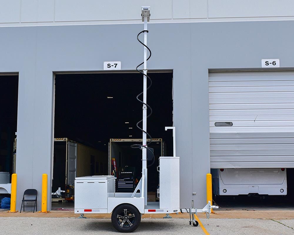 A white mobile sensing trailer with a vertical mast and equipment boxes, serving as a first responder unit in front of a garage