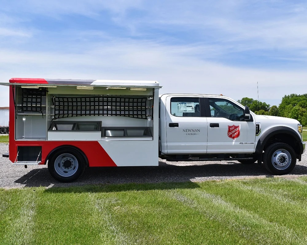 External view of a white Salvation Army Rapid Response Unit truck with red and white graphics, open side compartments revealing shelving and a serving area.