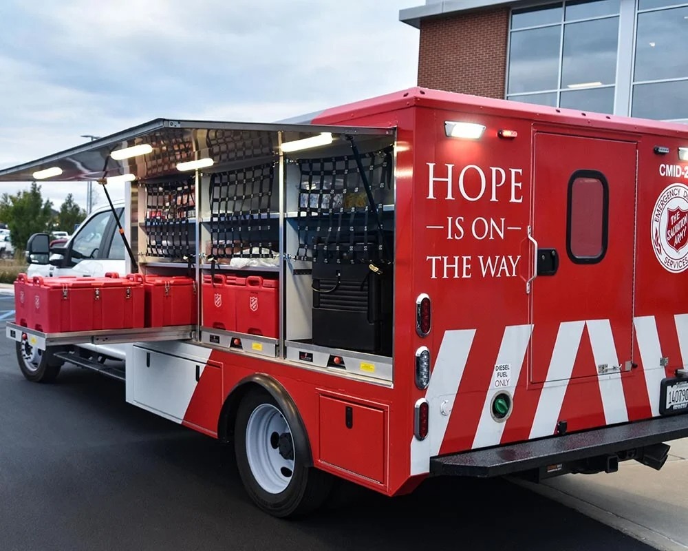 External view of a red and white Salvation Army Rapid Response Unit truck with the text HOPE IS ON THE WAY and open side compartments with storage bins.