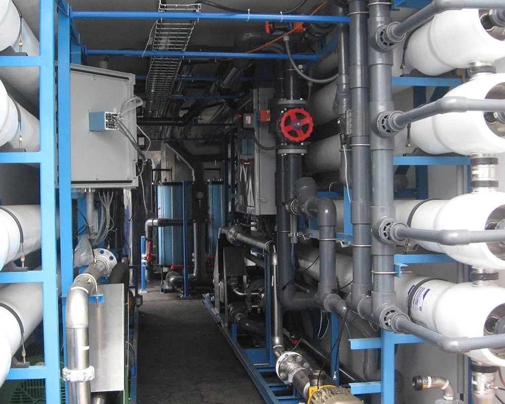 Interior view of an industrial fabrication water treatment unit housed in a shipping container, showing pipes, gauges, and white membrane vessels.