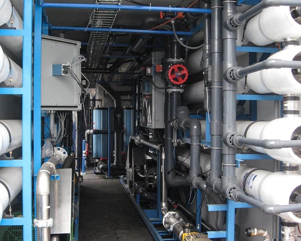 Complex Water Treatment Unit housed in a shipping container, showing white pressure vessels, blue structural frames, and gray PVC piping.